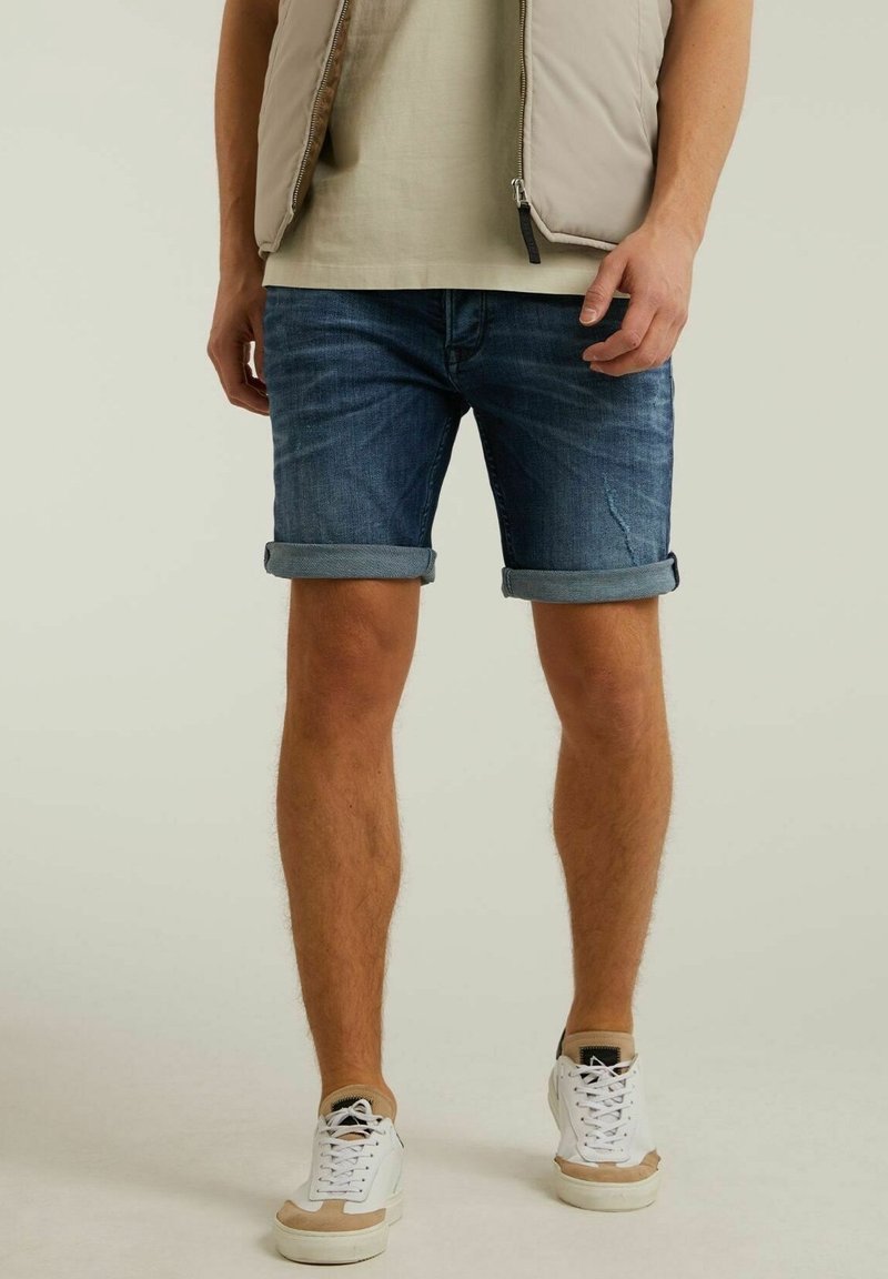 Shorts de mezclilla en azul oscuro, con dobladillos enrollados; combinados con zapatillas blancas y beiges. El modelo lleva un chaleco beige claro y una camiseta debajo.