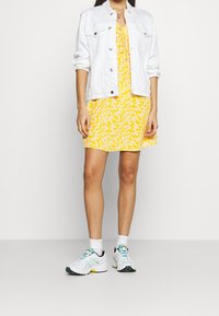 Valkoinen farkkutakki keltaisen kukkamekon päällä; kengät ovat valkoiset, joissa on turkooseja ja keltaisia yksityiskohtia, ja mukana on valkoiset sukat.