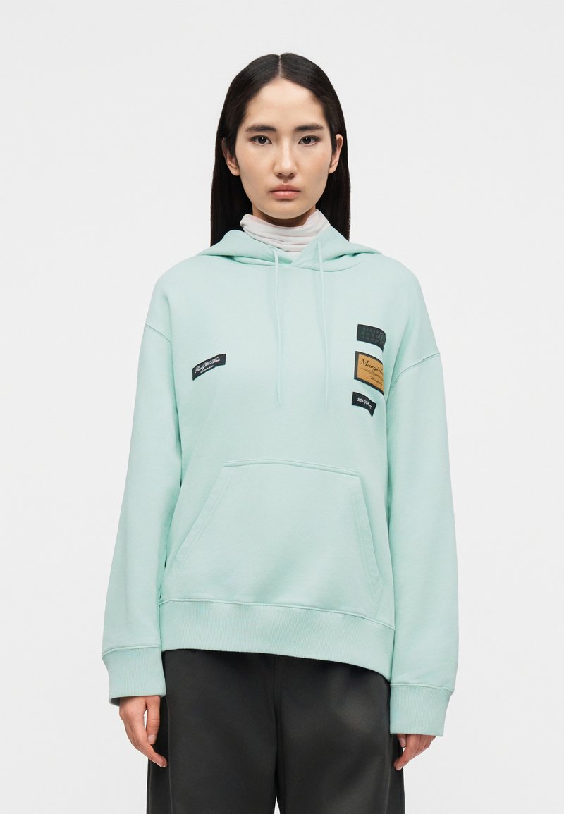 Γυναίκα με ίσια σκούρα μαλλιά φορά ει ανοιχτό μπλε φούτερ μετζή μπροστά και μπαλώματα, μαύρο παντελόνι και λευκό κοντό λαιμόκοψη από μέσα, ουδέτερο φόντο.