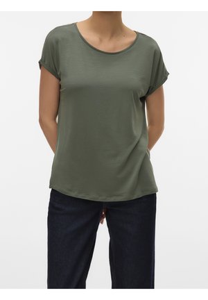 Persoon die een olijfgroen T-shirt met korte mouwen en een donkerblauwe spijkerbroek draagt, staand met handen achter de rug.
