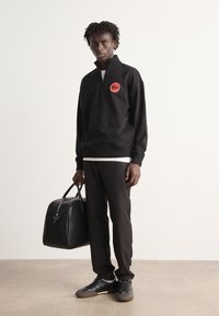 Sudadera negra con cremallera y logo rojo, pantalones negros y zapatillas negras. Sosteniendo un bolso de cuero negro. Diseño minimalista y colores neutros.