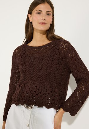 Femme portant un pull à maille ajourée marron foncé avec un ourlet festonné et un pantalon blanc, debout avec une main sur la hanche.