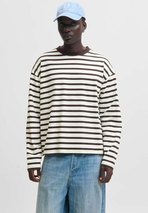 Personne portant une chemise blanche à manches longues avec des rayures horizontales marron, un jean en denim bleu clair et une casquette de baseball bleu clair sur un fond uni.