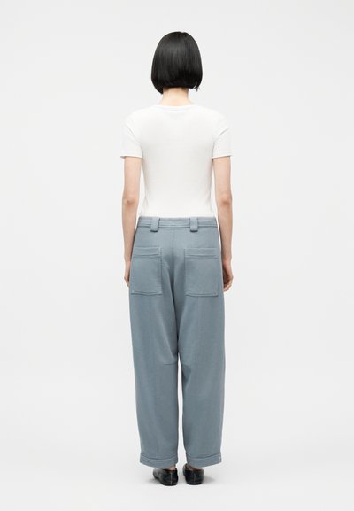 Άτομο με κοντά μαύρα μαλλιά στέκεται με την πλάτη, φοράει εφαρμοστή λευκή κοντομάνικη μπλούζα, φαρδιά ανοιχτομπλε παντελόνια με πίσω τσέπες και μαύρα παπούτσια.