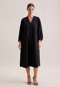 Schwarzes Midikleid mit V-Ausschnitt, locker sitzenden langen Ärmeln und glatter Textur. Der Saum reicht unter das Knie, kombiniert mit schwarzen, mit Riemchen versehenen High Heels.