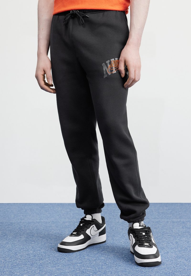 Person trägt schwarze Nike-Jogginghose mit orangefarbenem Logo am Oberschenkel, weiße Socken und schwarz-weiße Nike-Sneaker, steht auf blauem Boden.