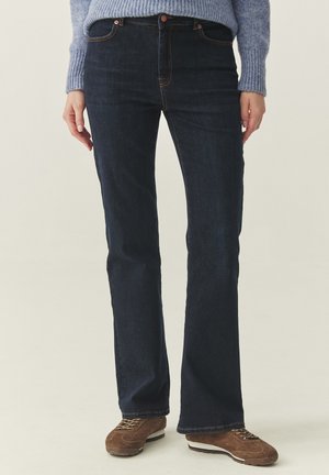 Osoba oblečená v tmavomodrých džínsach s rovnými nohavicami, svetlomodrom pletenom svetri a hnedých semišových teniskách, stojaca s uvoľnenými pažami po bokoch.