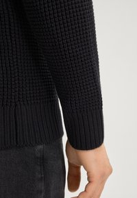 Marineblauwe gebreide trui met een gestructureerd patroon, geribbelde manchetten en een naadloos ontwerp dat overgaat in donkergrijze jeans.