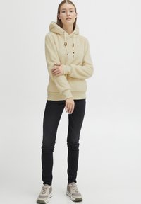 Beige fleecehuva med texturerad yta, justerbar dragsko och ribbade ärmslut. Matchas med svarta skinny jeans och ljusa sneakers.