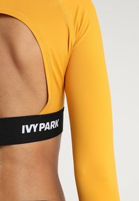 Gelbes Langarmoberteil mit offenem Rücken. Verfügt über ein schwarzes elastisches Band mit "IVYPARK"-Branding am Saum. Weicher, dehnbarer Stoff.