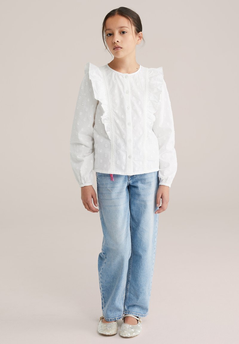 Blouse en coton blanc avec épaules à volants, motif floral texturé, boutons à l'avant, associée à un jean denim bleu clair et des chaussures argentées.