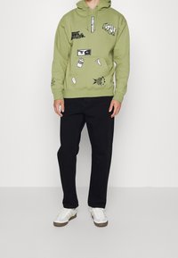 Felpa verde con grafiche stampate, dotata di cappuccio con coulisse, tasca a marsupio e polsini a costina. Abbinata a pantaloni neri e sneakers.