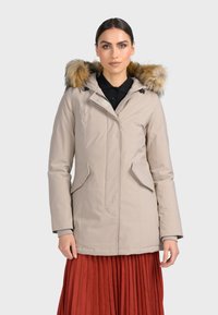 Beige zateplená bunda s kapucí podšitou umělou kožešinou, se zipem, bočními kapsami a elegantním designem. Nositá přes černý top a červenou sukni.