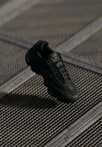 Basket noir sportif avec une tige texturée, des lacets et un col rembourré. Logo proéminent sur le côté, sur fond à motifs.