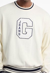 Suéter de punto color crema con un cuello redondo a rayas azul marino, puños acanalados y una gran "G" texturizada con "JEANS" debajo.