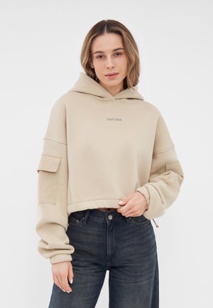 Felpa corta beige con cappuccio, maniche lunghe e zip laterali. Presenta un logo discreto sul petto e ampie tasche a toppa sulle maniche.