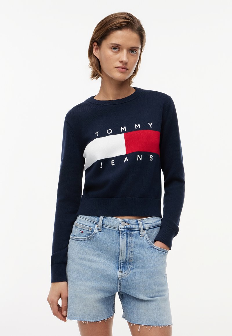 Γυναίκα που φοράει φούτερ Tommy Jeans σε μπλε ναυτικό χρώμα με κόκκινο και λευκό λογότυπο και ανοιχτόχρωμα τζιν σορτς, στέκεται μπροστά σε ένα απλό λευκό φόντο.