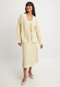 Stickad tvådelad outfit i krämfärg, bestående av en knapp-up cardigan och en matchande midikjol med vertikalt ribbad textur och en lätt lyster.