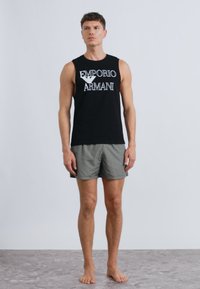 Μαύρο αμάνικο τοπ με το λογότυπο "Emporio Armani", σε συνδυασμό με γκρι ριγέ σορτς. Το μοντέλο στέκεται ξυπόλητο πάνω σε υφή επιφάνειας.