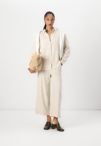 Helles beige Hemd mit Knöpfen, weite passende Hose und dunkle Absatzschuhe. Die Textur ist glatt; beinhaltet eine geflochtene Tasche.