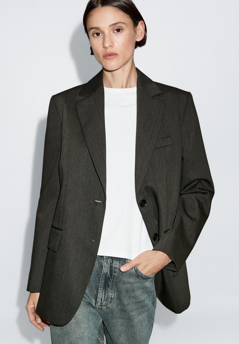 Donkergrijze op maat gemaakte blazer met een enkele knoopsluiting, notch-kraag en twee voorzakken, gedragen over een wit T-shirt en blauwe spijkerbroek.