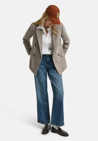 Blazer grigio doppiopetto sopra una camicia bianca, abbinato a jeans blu a gamba larga. Un cappellino arancione con il logo 'G' e mocassini marroni completano il look.