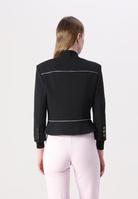Černý spřádaný sako s peplum lemem, zdobené bílým lemováním a zlatými knoflíky na rukávech. Hladká tkanina se strukturovanou siluetou.