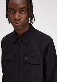 Giacca nera con zip e due tasche frontali, realizzata in materiale texturizzato. Presenta un piccolo logo ricamato verde sulla tasca sinistra.