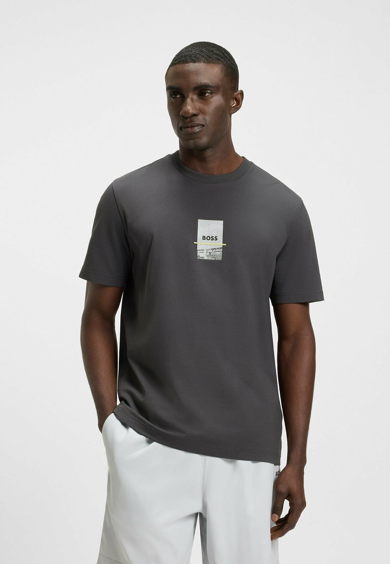 Camiseta gris de manga corta con cuello redondo, que presenta un logo impreso y acentos gráficos en la parte frontal. Confeccionada en tejido suave y ligero.