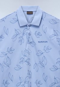 Ljusblå polo med broderade lövmönster, tre knappar och klassisk krage. Har "NAPAPIJRI"-logotyp i matchande tråd.