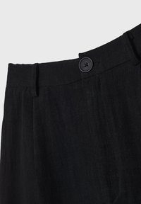 Gros plan de la ceinture d'un pantalon en tissu noir avec un bouton noir fermé et une boucle de ceinture sur le côté.