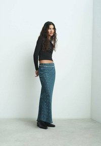 Larga falda de color teal con un estampado floral y un sutil brillo, combinada con un top corto negro y botas de tobillo negras. Silueta suave y ajustada.