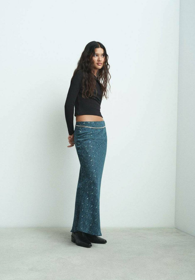 Lång teal-färgad kjol med blommönster och subtil lyster, kombinerad med en svart crop top och svarta ankelstövlar. Slät, figurnära silhuett.