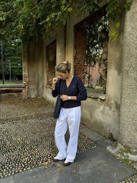 Marinblå knäpp-up topp, vita vida byxor och bruna skor, mot en texturerad stenstig och en delvis raserad vägg med grönska.