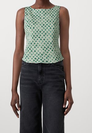 Haut sans manches à motif géométrique vert et blanc porté avec un jean noir taille haute sur une personne debout devant un fond uni.