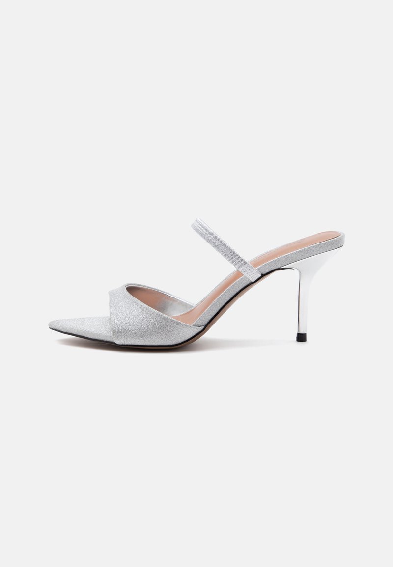 Mule argentée à talon haut avec bout pointu, large bande pailletée sur les orteils et fine bride sur le dessus du pied, sur fond blanc.