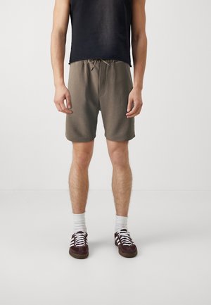 Homme portant un short vert olive à cordon, des chaussettes blanches montantes, des baskets marron foncé et un débardeur noir, debout sur un fond clair.