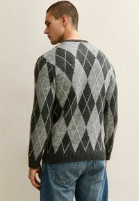 Man staat naar links gekeerd, gekleed in een donker en licht grijs ruitjestrui en een blauwe spijkerbroek, tegen een effen beige muur.