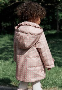 Veste matelassée à carreaux roses et blancs avec capuche, dotée d'un col à volants, portée avec des leggings clairs et des bottes en fourrure, en extérieur.