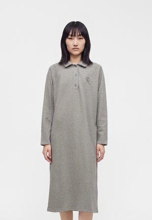 Donna con capelli neri lisci, indossa un vestito grigio a maniche lunghe, lungo fino al ginocchio, con un piccolo emblema del sole sul petto, in piedi davanti a uno sfondo bianco.