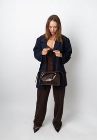 Geantă de mână din piele maro, cu design plisat și închidere cu șnur. Modelul poartă un blazer supradimensionat închis la culoare și pantaloni asortați.