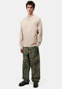 Sudadera beige con puños acanalados, combinada con pantalones holgados de camuflaje en verde y marrón, completada con zapatos negros. Detalle de cadena plateada.