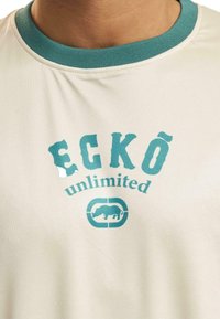 Kremfarget T-skjorte med turkise ribbekanter og grafisk trykk. Teksten sier "ECKO unlimited" med et neshornikon, mykt stoff, avslappet passform.