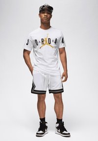 Balts t-krekls ar melnu un zelta "JORDAN" grafiku, pārī ar baltām šortiem ar melnu apmali un akcentiem, papildināts ar melnbaltiem sporta apaviem.