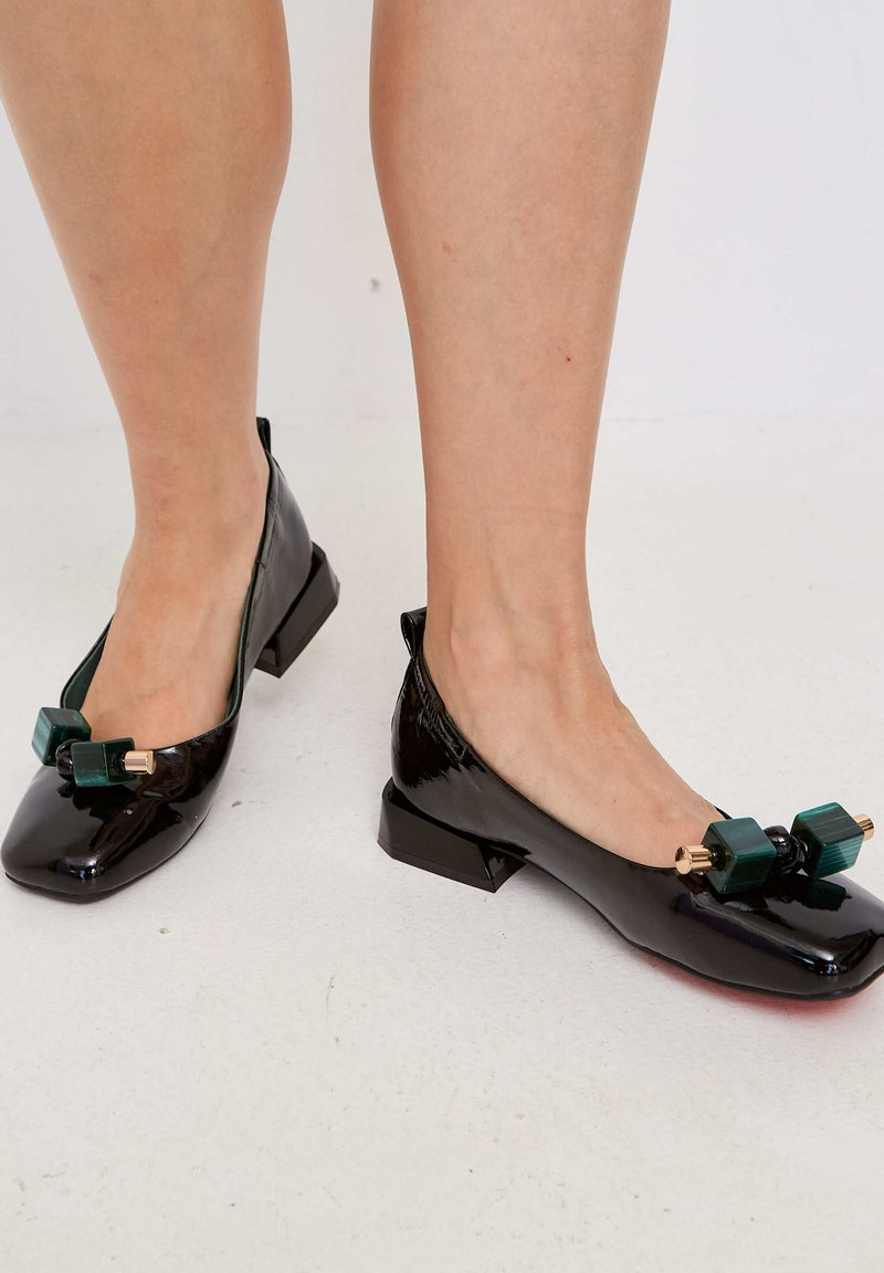 Ballerines en cuir verni noir avec un orteil carré, présentant des accents décoratifs verts et un petit talon épais. Texture lisse dans l'ensemble.