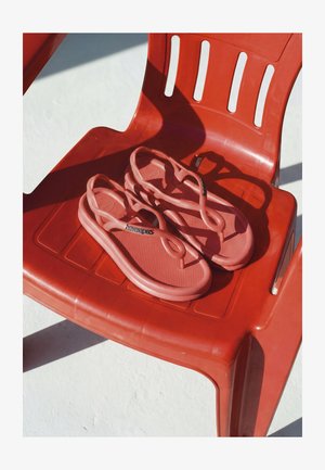 Paire de sandales Havaianas rose corail reposant sur une chaise en plastique rouge projetant des ombres sur une surface blanche.