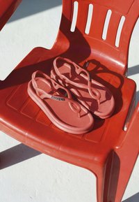 Paire de sandales Havaianas rose corail reposant sur une chaise en plastique rouge projetant des ombres sur une surface blanche.