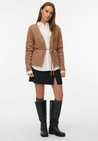 Rust boja pleteni kardigan s remenom, nosi se preko bijele košulje s gumbima, uparen s crnom suknjom i crnim čizmama do koljena.