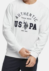 Bílá mikina s ribovanými manžetami a lemem, která má námořnickou grafiku s jezdeckými hráči a textem "AUTENTICKÝ US PA" v tučném písmu.