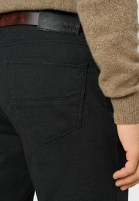 Zwarte textuurbroek met een zichtbare achterzak, leren label "BRAX" en een lichtbruine trui die gedeeltelijk de taille bedekt.